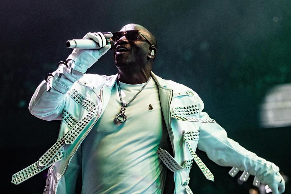 Akon performs on stage in a white studded jacket, sunglasses, and a pendant necklace.