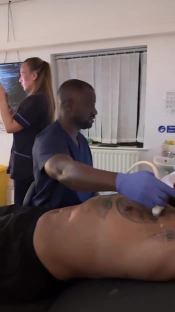 A man in blue scrubs performs an ultrasound on a patient lying on an examination table, while a woman watches a screen in the background.