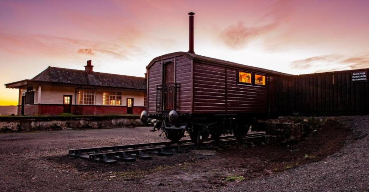 A converted sleeper carriage sits on tracks in front of a station house at sunset.