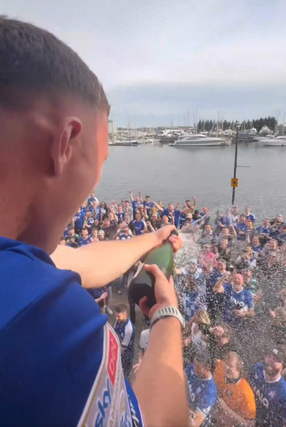 Man in a blue shirt spraying champagne over a crowd of cheering people, with a marina in the background.