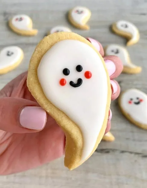 A person holding up a decorated ghost-shaped Halloween cookie with white icing, black eyes and mouth, and red cheeks, with other similar cookies in the background.