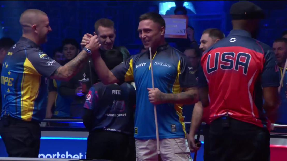Gerwyn Price in a blue and yellow shirt, holding a pool cue, celebrating with another player, with other players in the background.