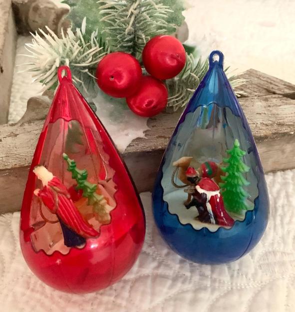 Two Jewel Brite Christmas ornaments, red and blue, with nativity scenes inside, set against a frosted pine branch and red berries.