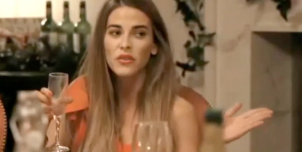 Woman in an orange top holding a glass, gesturing with her other hand.