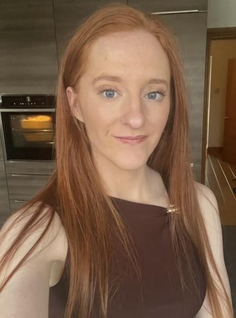 A woman with long red hair and blue eyes smiles at the camera while baking a dessert in the oven.