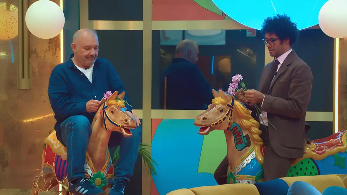 Bob Mortimer and Richard Ayoade sitting on carousel horses with flowers.