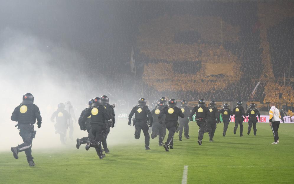 Police in riot gear run on a foggy soccer field with a crowd in the background.