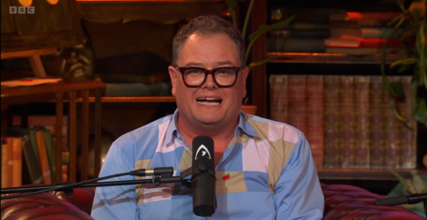 Alan Carr sitting in front of a microphone, wearing a patterned shirt and glasses.