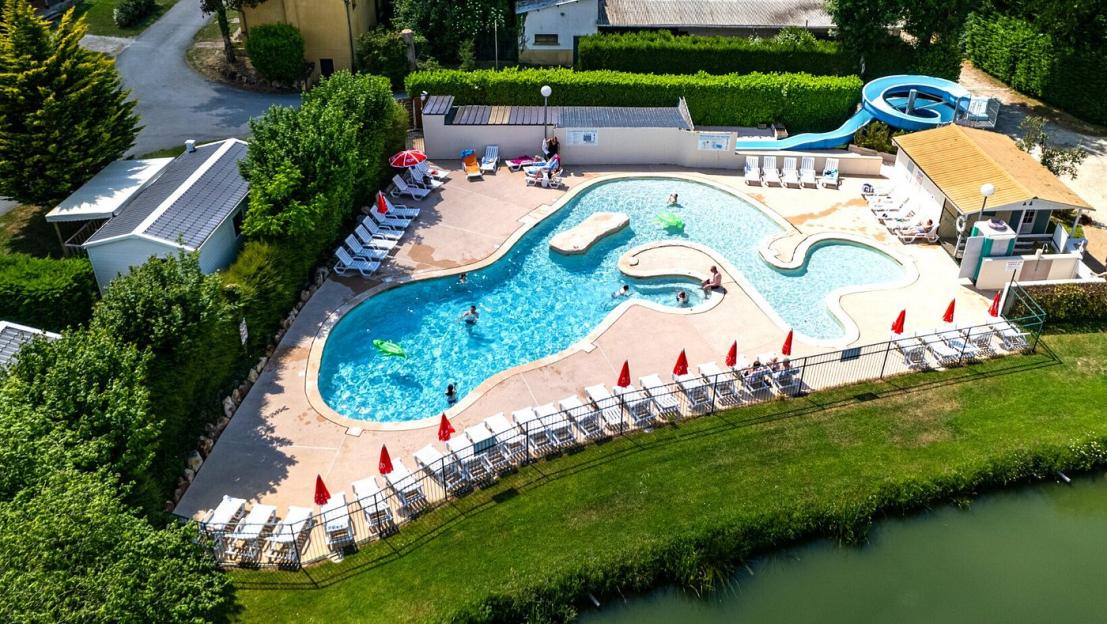 Aerial view of a swimming pool complex with a curvy pool, lounge chairs, red umbrellas, a waterslide, and surrounding buildings and greenery next to a body of water.