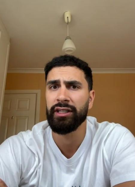 A man with a beard and dark hair, wearing a white t-shirt, looking at the camera with a confused expression.