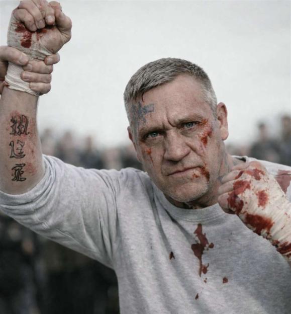 A male strongman with blood on his face, bandages, and shirt, raising a fist with tattoos on his arm.