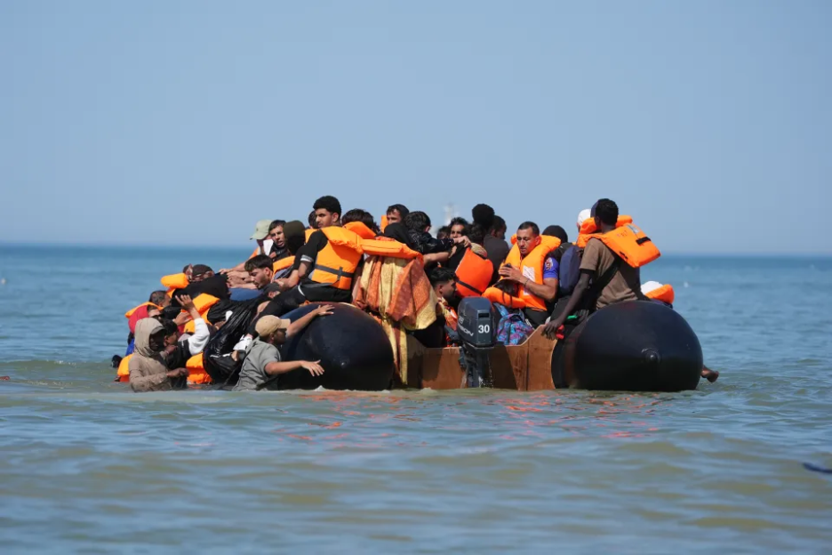 An overcrowded boat filled with migrants wearing orange life vests on the sea.