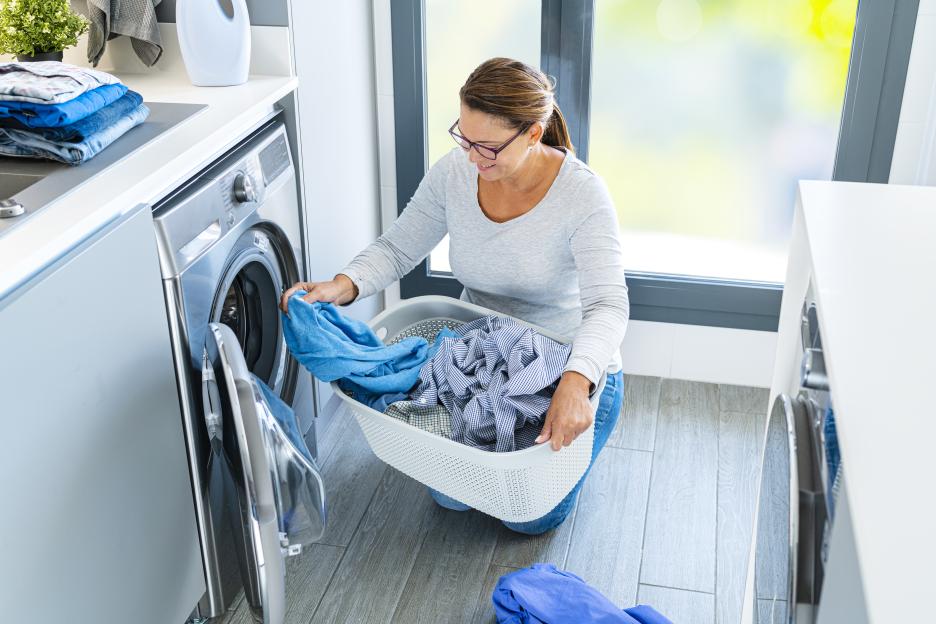 A woman loads clothes from a laundry basket into a front-loading washing machine.