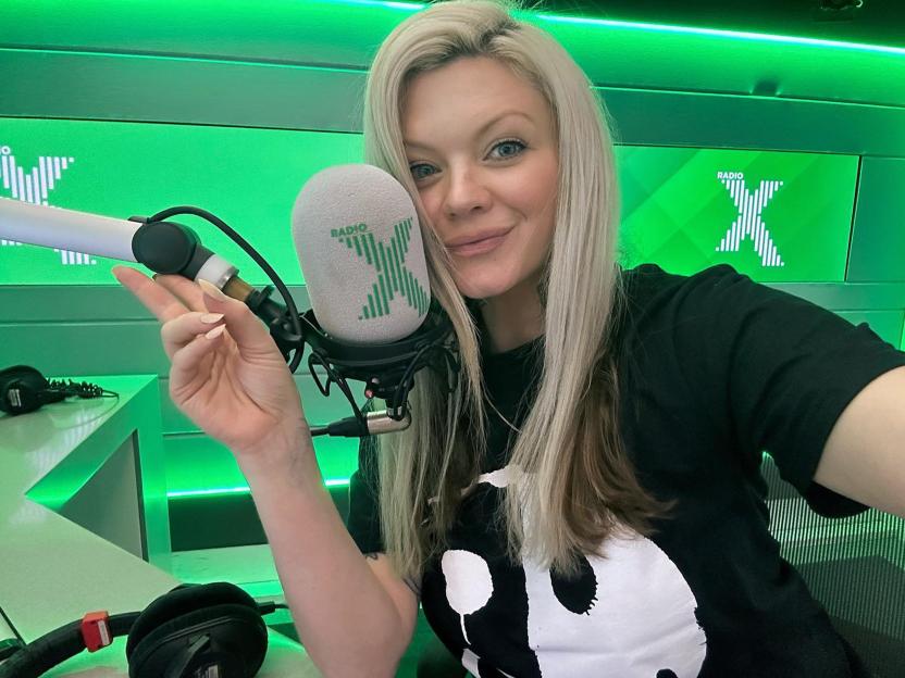 Polly James, a radio presenter, smiling in a radio studio with a microphone next to her face and the Radio X logo visible in the background.