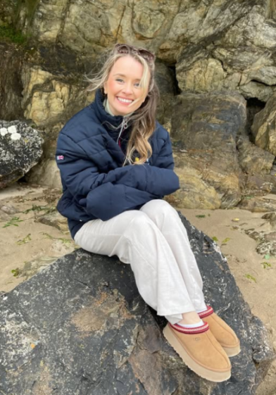 A woman smiling at the camera while sitting on a rock in a dark blue puffer jacket, white pants, and brown Uggs with a red and white stripe.