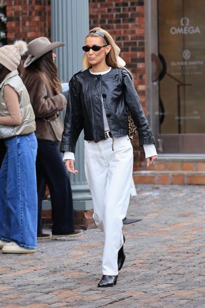 Bella Hadid in a black leather jacket, white pants, and black sunglasses, walking on a cobbled street.
