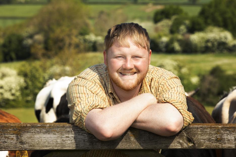 Kaleb Cooper smiling at the camera, resting his crossed arms on a wooden fence with cows in the background.