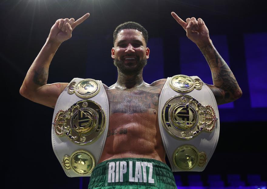 Ty Mitchell, a boxer, smiles while wearing two championship belts and pointing to the sky.