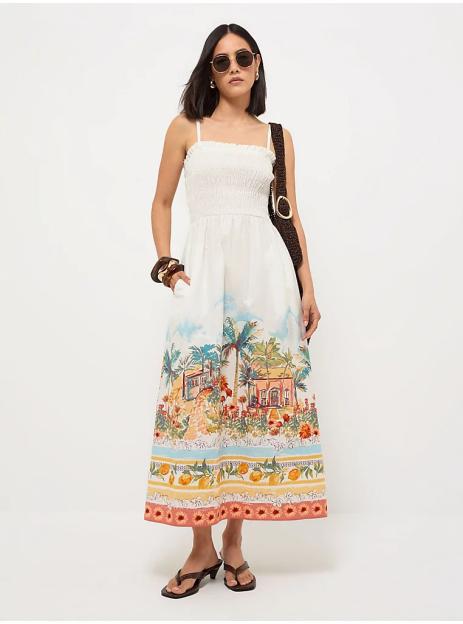 A woman in a white shirred midi dress with a colorful scenic tropical print, dark sunglasses, and flip-flops.