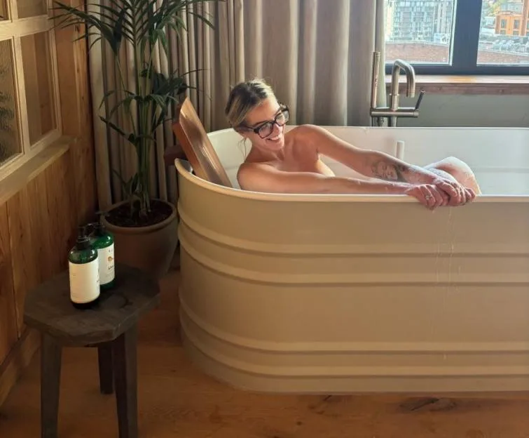Helen Flanagan smiling while sitting in a bathtub by a large window.