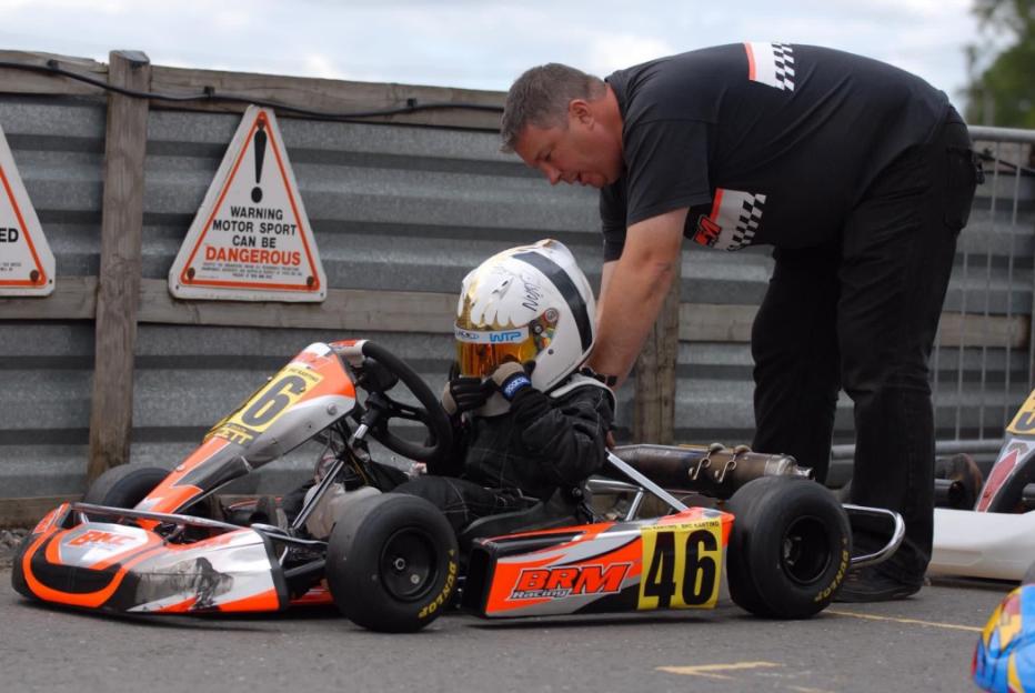 A man leans over to assist a young child in a go-kart, with a "WARNING MOTOR SPORT CAN BE DANGEROUS" sign in the background.