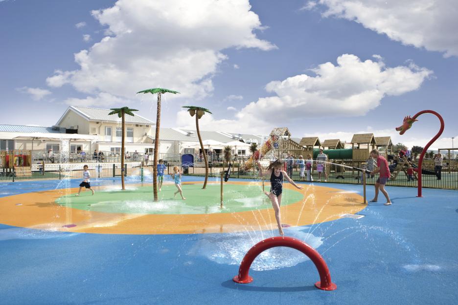 Children playing in a water park with palm tree sprinklers.
