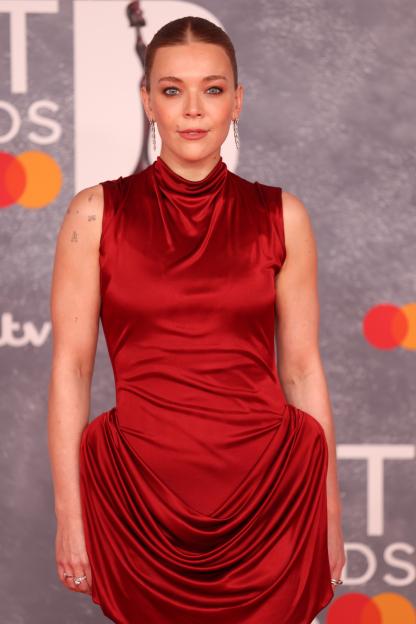 A woman in a red satin dress with a high neckline and draping at the hips, wearing dangling earrings and a ring, poses at the BRIT Awards 2025 arrivals.