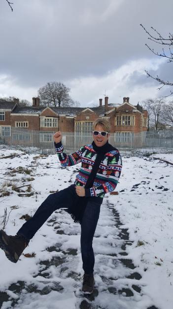 Hollywood filmmaker Hopwood DePree in a Christmas sweater, black scarf, and white sunglasses, poses in a snowy field with a brick building in the background.
