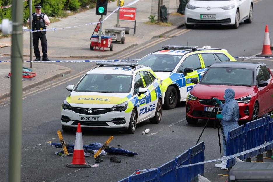 Forensic team member taking photographs of a crime scene with police cars and barricades.
