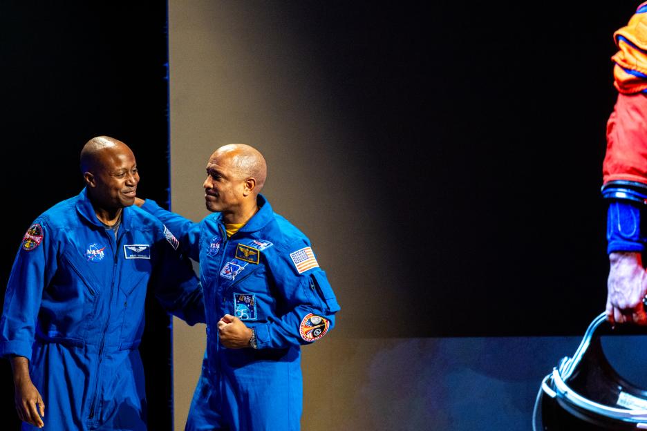 Two Artemis III crew members, Andre Douglas and Victor Glover, stand on a stage in their blue flight suits.