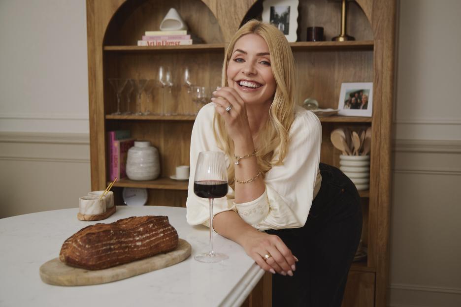 Holly Willoughby smiling while leaning on a counter with a glass of red wine and a loaf of bread.