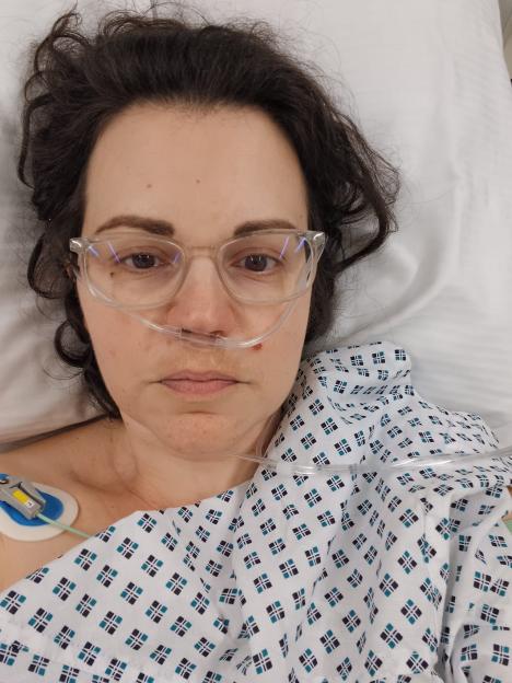 Dr. Philippa Kaye in a hospital gown, wearing glasses and a nasal cannula, lying in bed after a huge operation for bowel cancer.