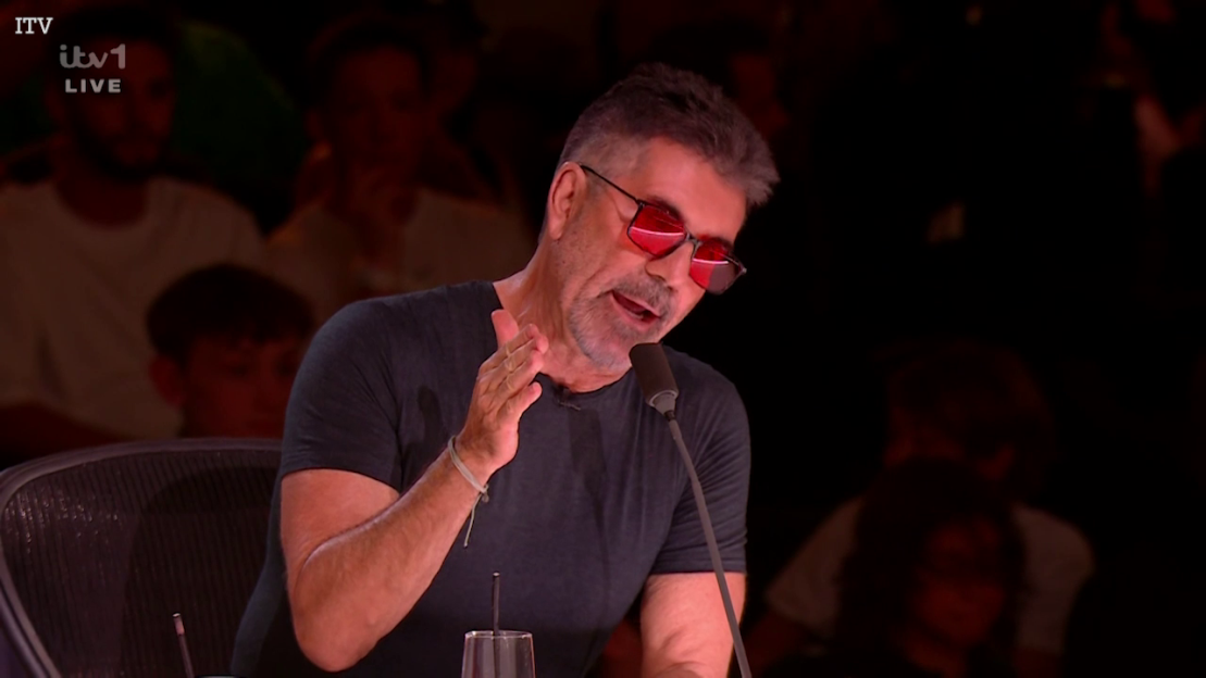 Simon Cowell in red-tinted sunglasses speaking into a microphone.