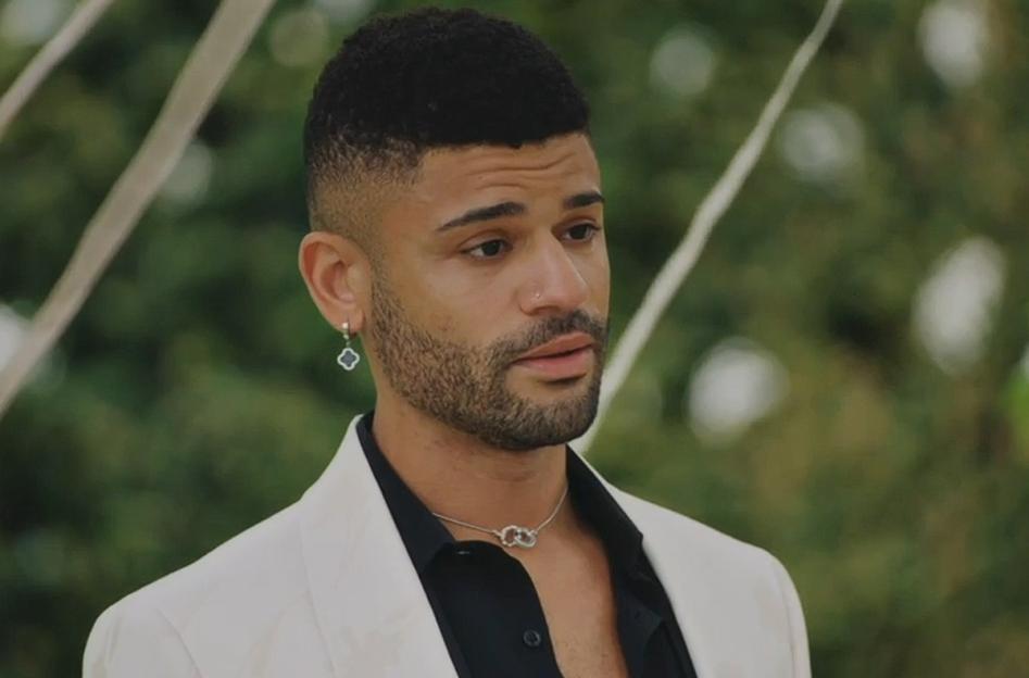 A tearful man in a white blazer and black shirt, with a clover-shaped earring, looking distressed.
