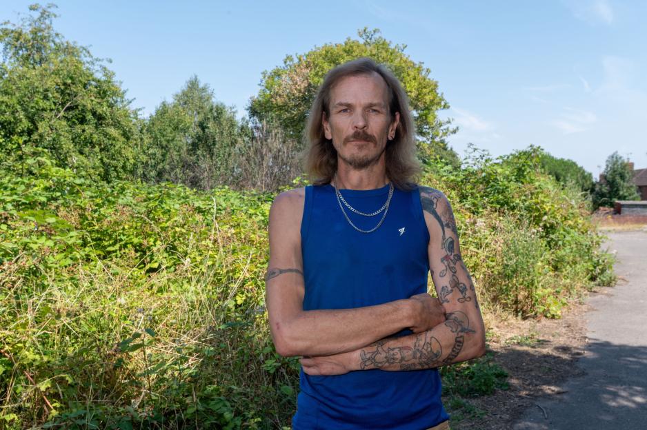 Gary Berresford in a blue tank top, with long hair and a mustache, standing in front of lush green bushes and trees.