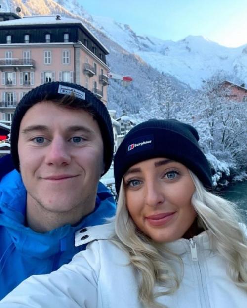 Adam Muskett and Katie Worrell in winter clothing, posing together with a snowy mountain landscape and buildings in the background.