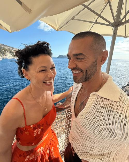 Emma and Matt Willis laughing under an umbrella by the sea.
