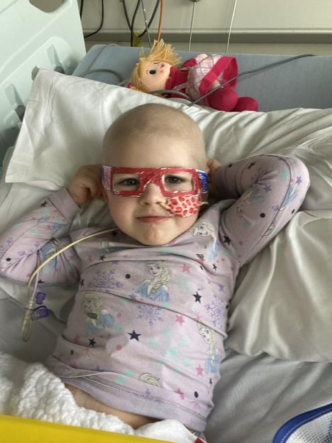 Poppy Catterall, a child with Acute Lymphoblastic Leukaemia, lying in a hospital bed with a doll, wearing red glasses and a Frozen pajama set.