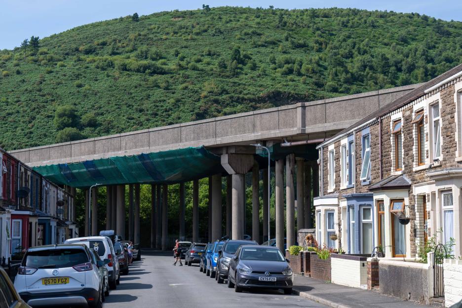 Row of terraced houses on a street running under an elevated concrete motorway, with a large green hill in the background.