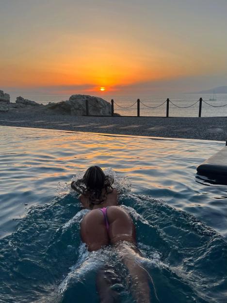 Woman in a pink bikini swimming in a pool towards the ocean at sunset.