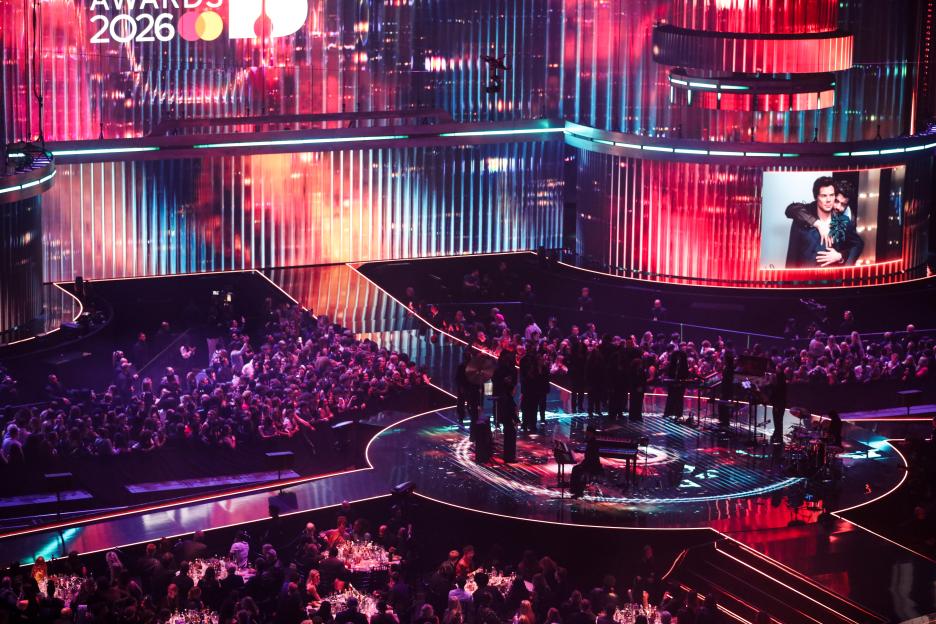 General view of the stage at The BRIT Awards 2026, showing the audience, an orchestra, and large screens displaying colorful lights and an image of two men.