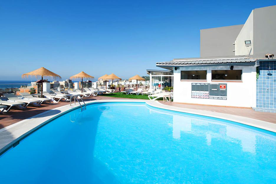 Rooftop swimming pool with lounge chairs and straw umbrellas, overlooking the ocean.