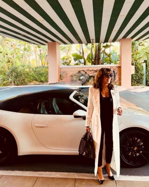 A woman in a white coat and black outfit stands next to a white Porsche, holding a patterned bag.