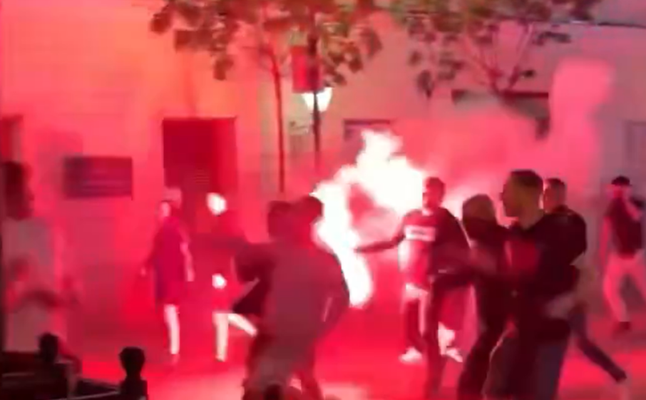 A crowd of men in a street illuminated by bright red light and smoke from a flare.