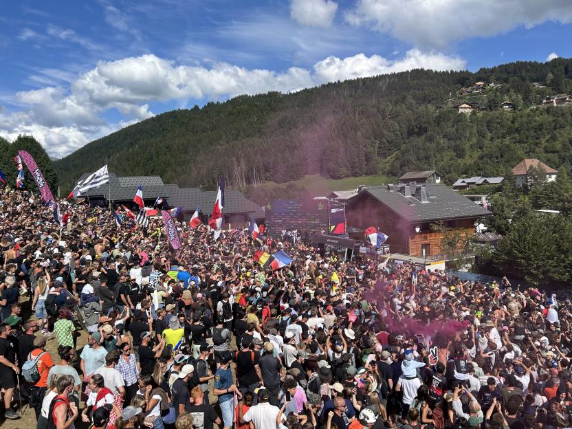 Large crowd of people gathered in a mountainous area with trees and buildings, some holding flags and pink smoke visible.