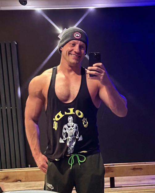 A man with a muscular physique wearing a black Gold's Gym tank top and a grey beanie smiles while taking a mirror selfie.