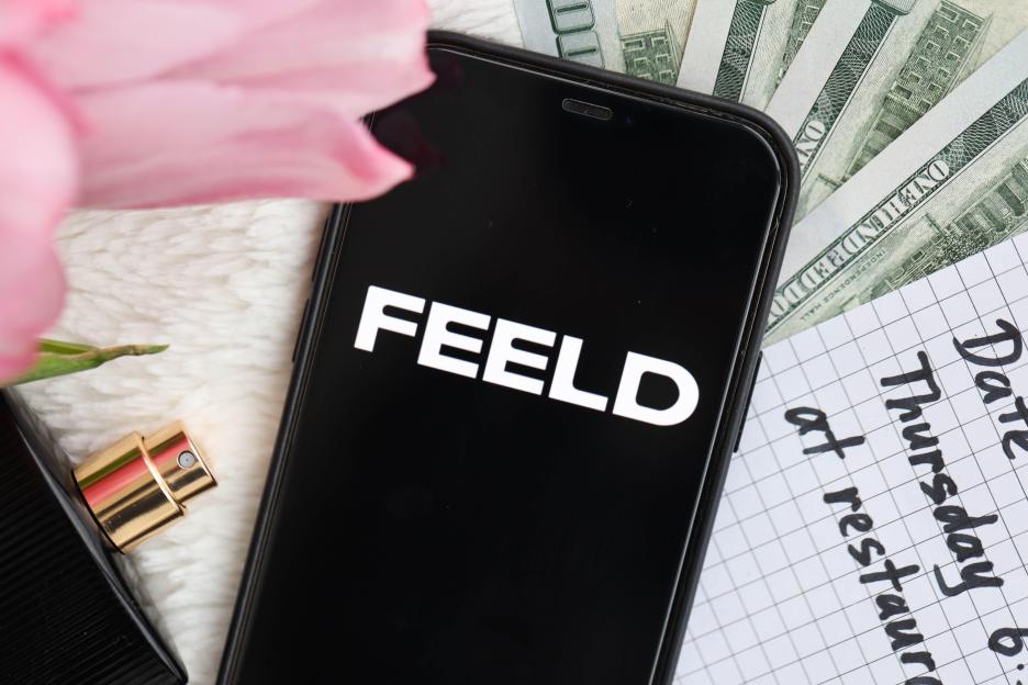 Close-up of an iPhone displaying the Feeld dating app logo, alongside perfume, cash, and a note reading "Date Thursday at restaurant."