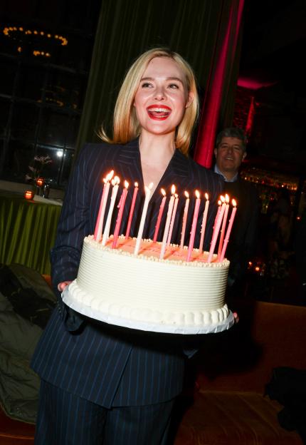 Elle Fanning smiles while holding a large birthday cake with lit candles.