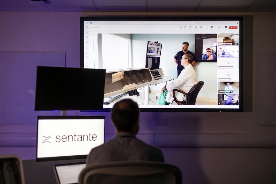 A person viewing a large screen displaying a video conference with a team of surgeons, one of whom is operating a machine.