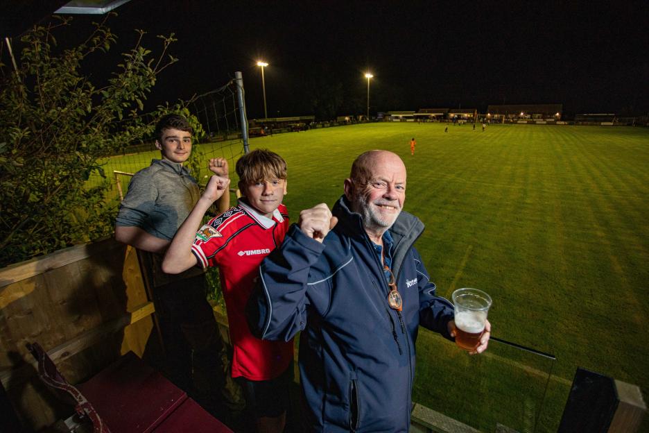 Three football fans, a man and two boys, cheer from a homemade stand overlooking a brightly lit football field at night.